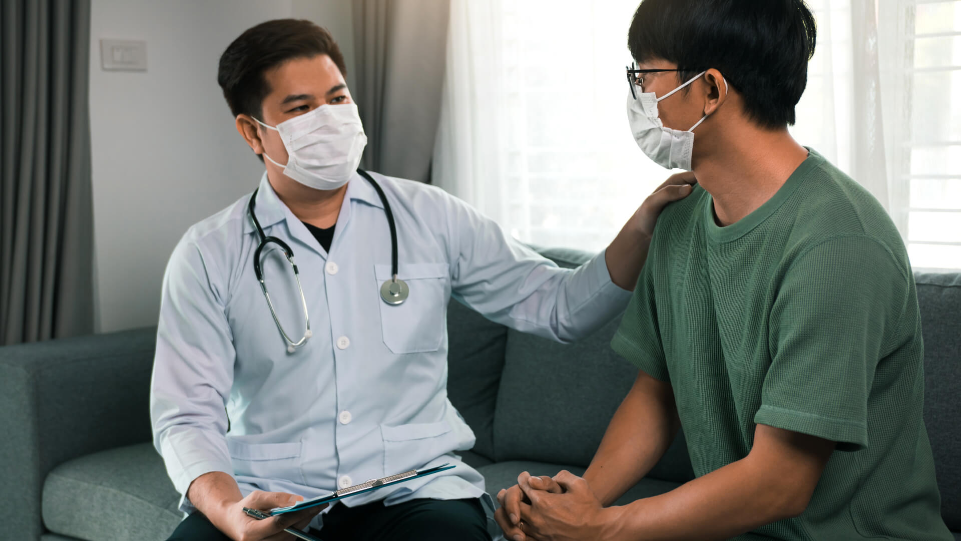 Doctor in a face mask with a stethoscope comforting a patient, also in a mask, during a consultation in a home setting, emphasizing personal care in healthcare.