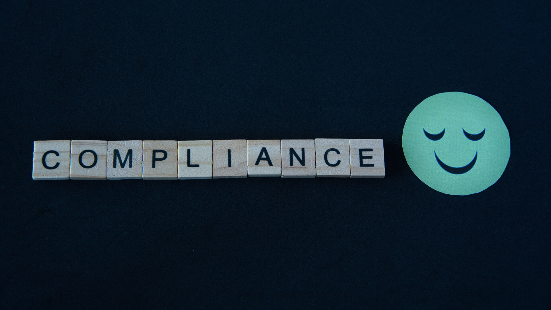 Scrabble letters on a dark background spelling out 'COMPLIANCE' next to a paper smiley face, symbolizing the positive aspects of adhering to rules and regulations.