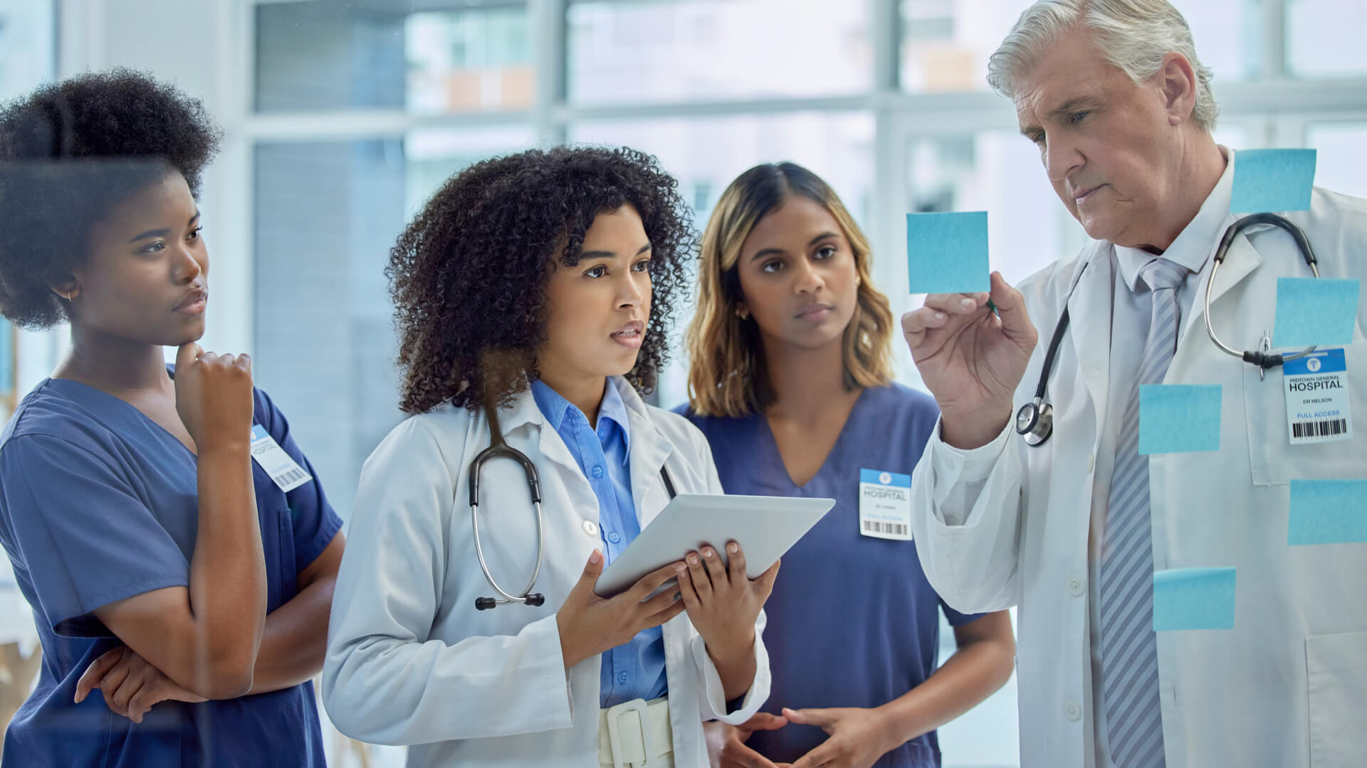 A diverse team of healthcare professionals in a clinical setting, with a senior doctor holding up sticky notes during a collaborative discussion, while a female doctor with a tablet listens intently, illustrating teamwork and communication in patient care coordination.