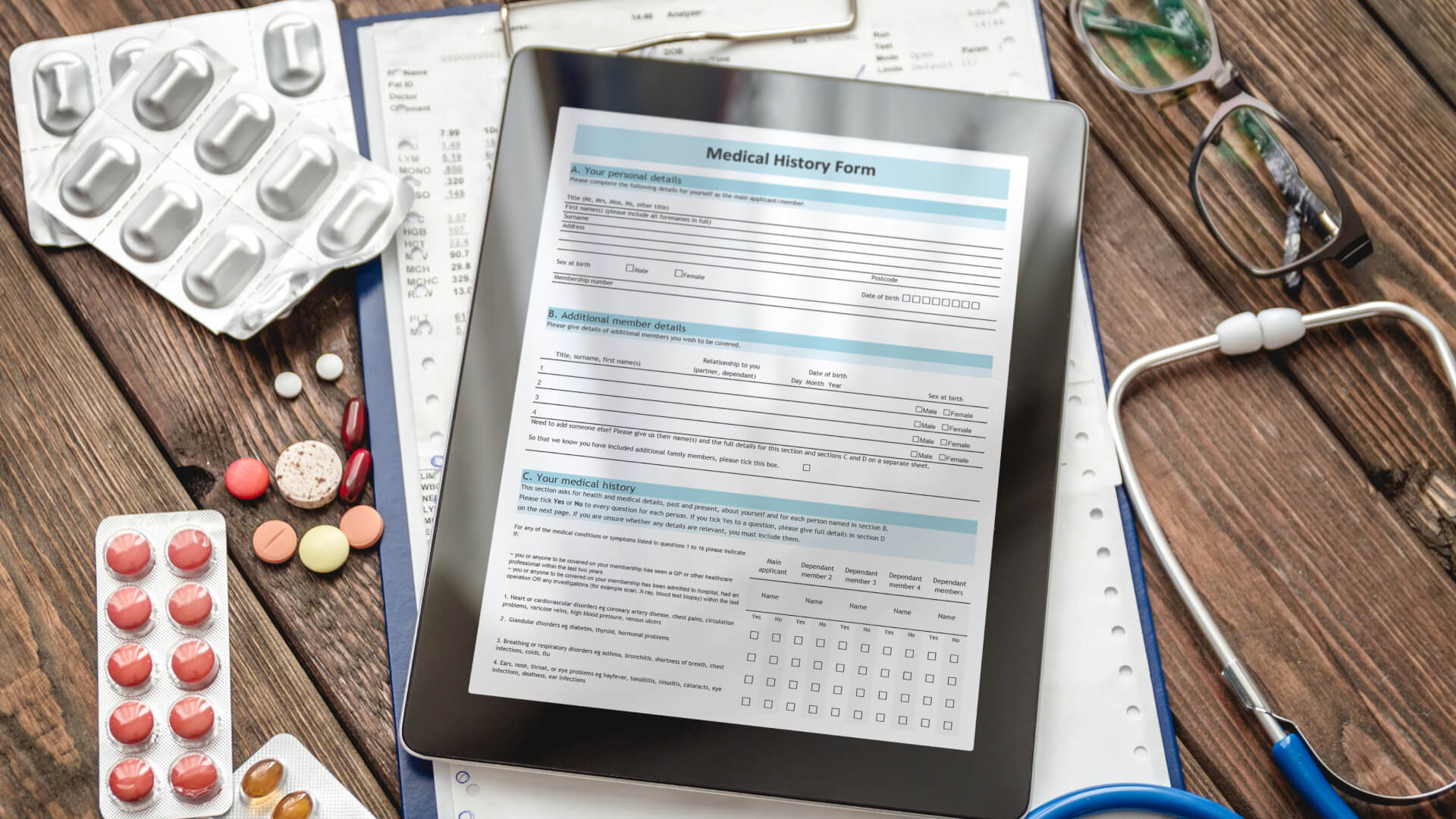 A digital tablet displays a Medical History Form, resting on a wooden table alongside a stethoscope, eyeglasses, and various medication packs, depicting the convenience and organization of electronic medical records in healthcare management.