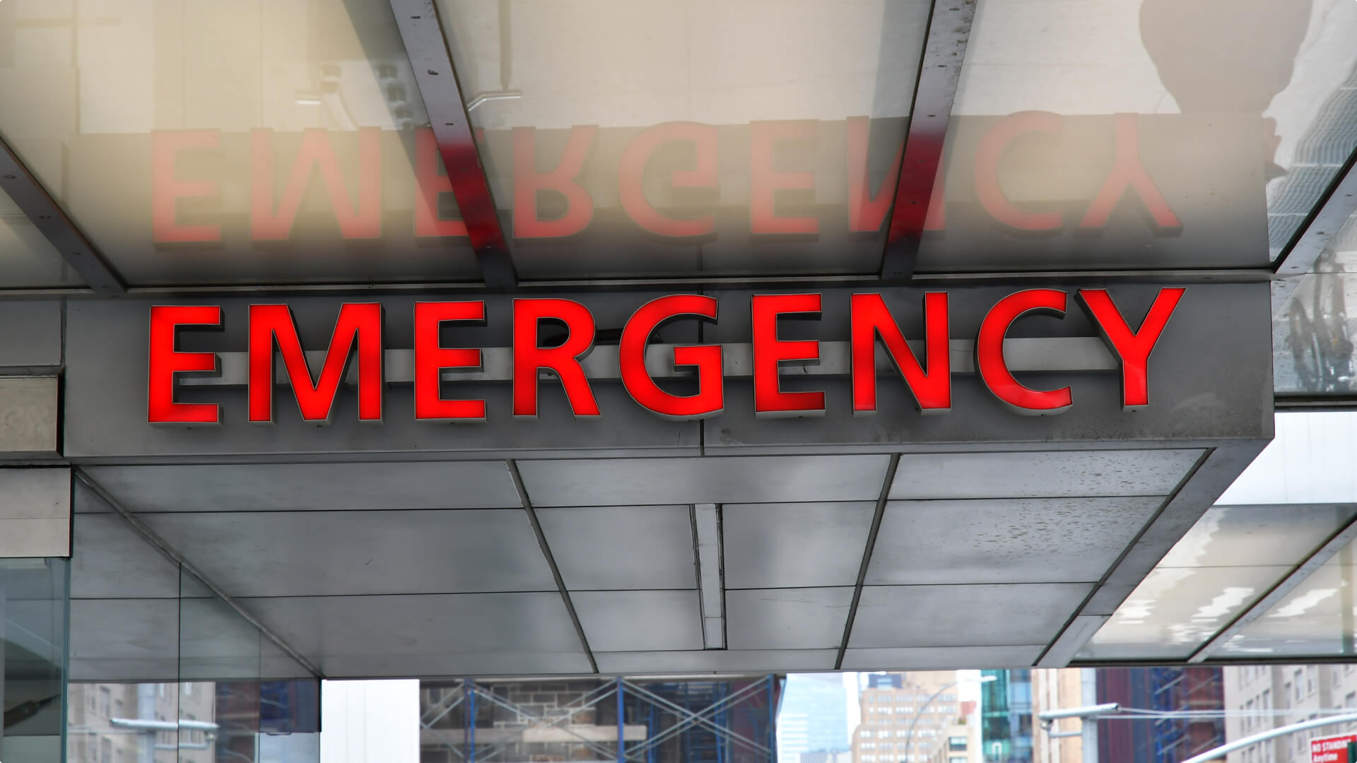 The word 'EMERGENCY' in bold red letters is affixed to the facade of a building, likely indicating the entrance to an emergency room or department, with its reflection visible on a glass surface above.