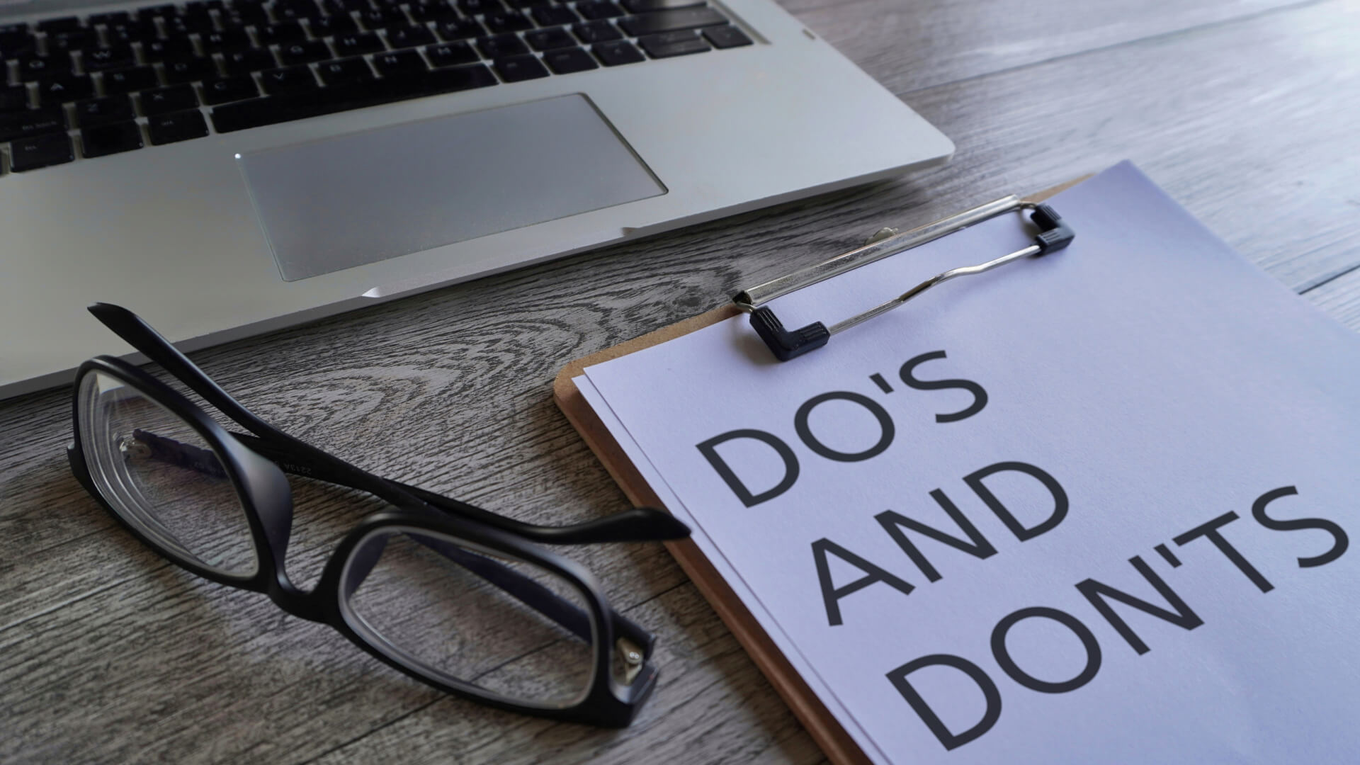 A laptop keyboard, a pair of eyeglasses, and a clipboard with the text DO's AND DON'Ts on a wooden surface.