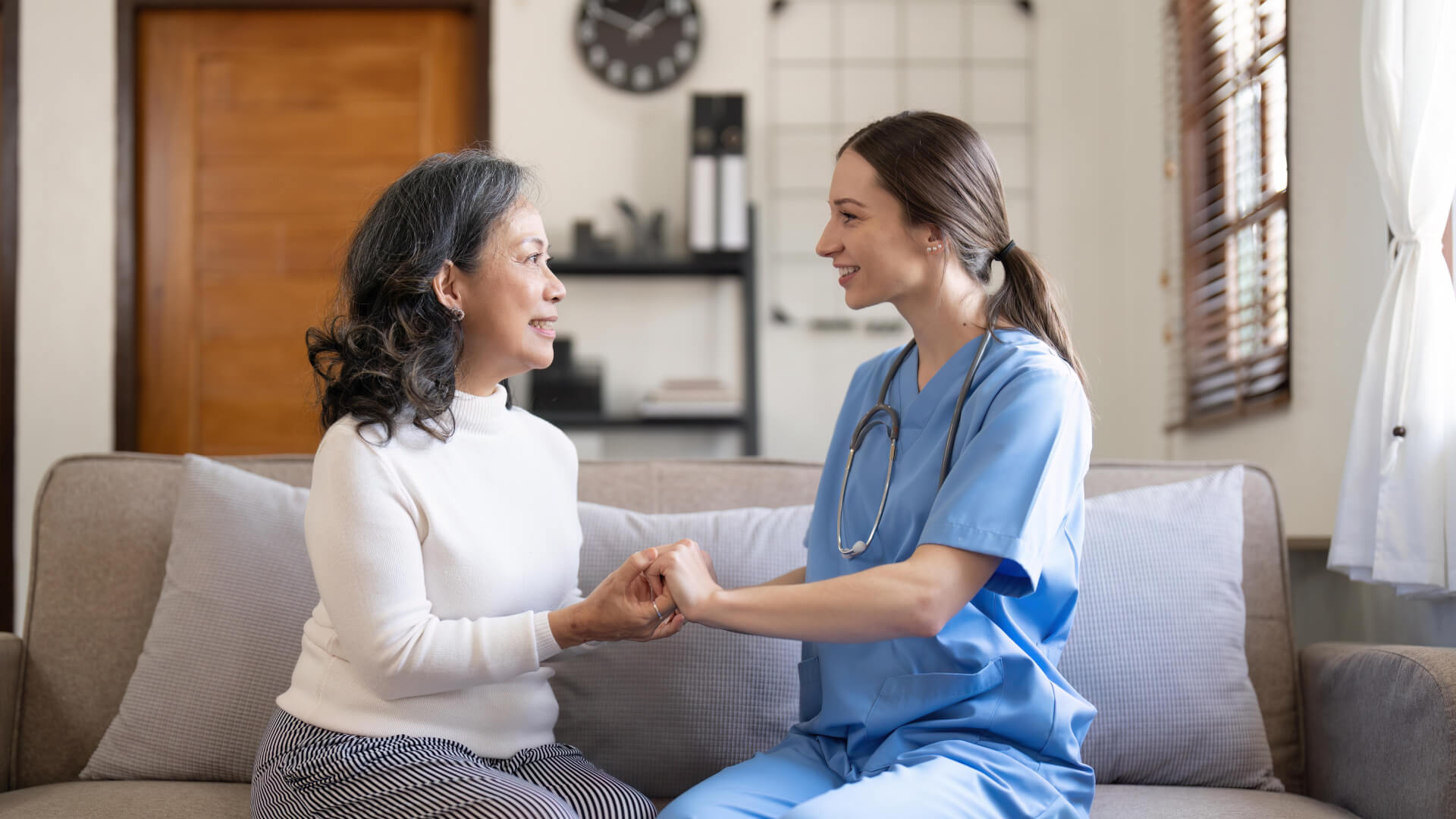 A healthcare professional in blue scrubs holding hands with an elderly patient, showing a comforting interaction in a home setting.