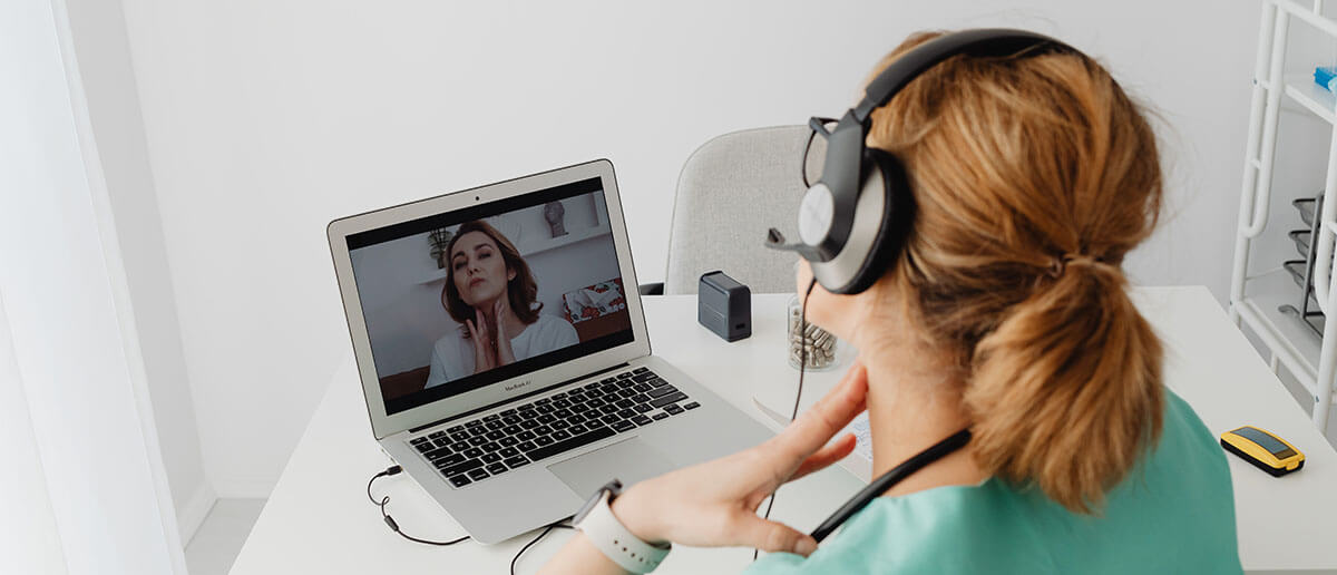 a doctor wearing headphones doing a video call with a patient on her laptop