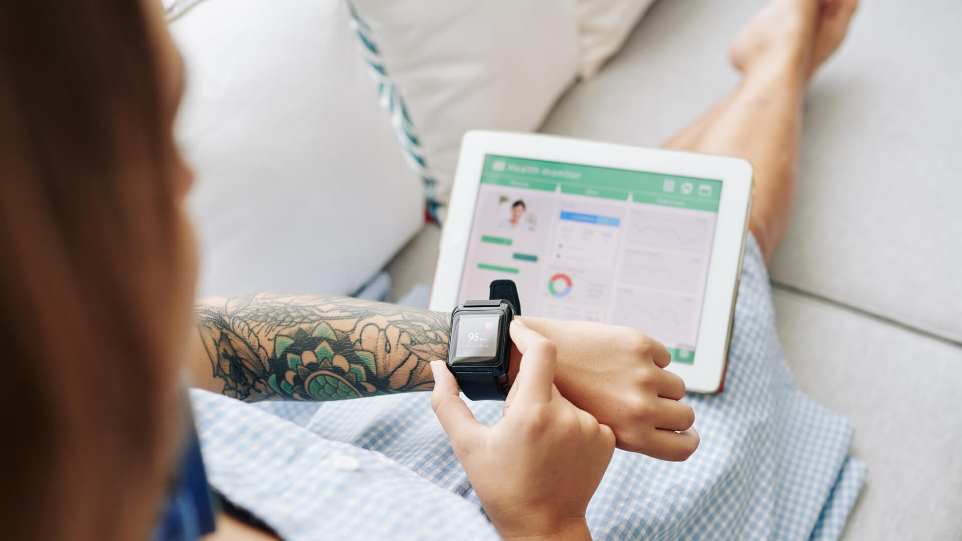 a woman is sitting on a couch with a tattooed arm using her smartwatch with a tablet placed on her lap